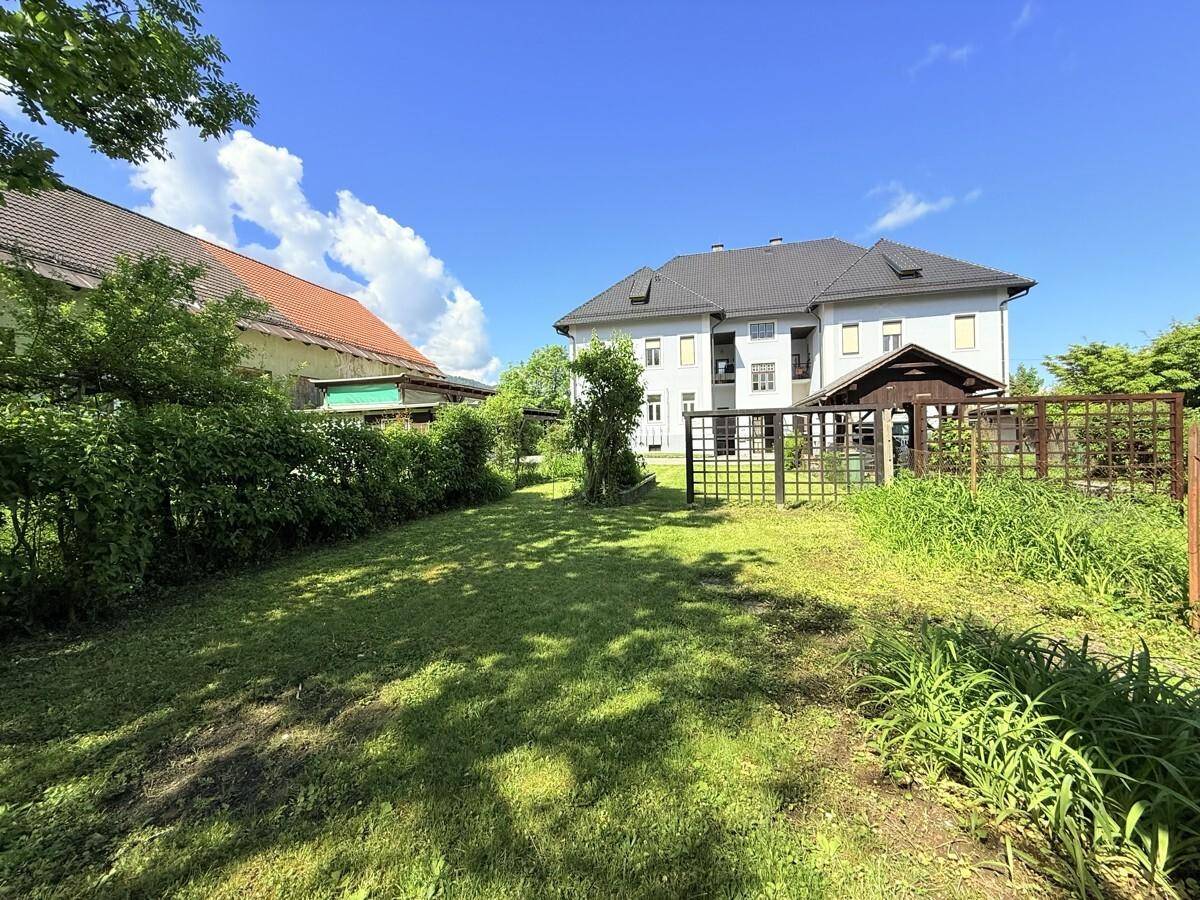 Weitläufiger Garten mit Rasenfläche und einem Mehrfamilienhaus im Hintergrund unter blauem Himmel.