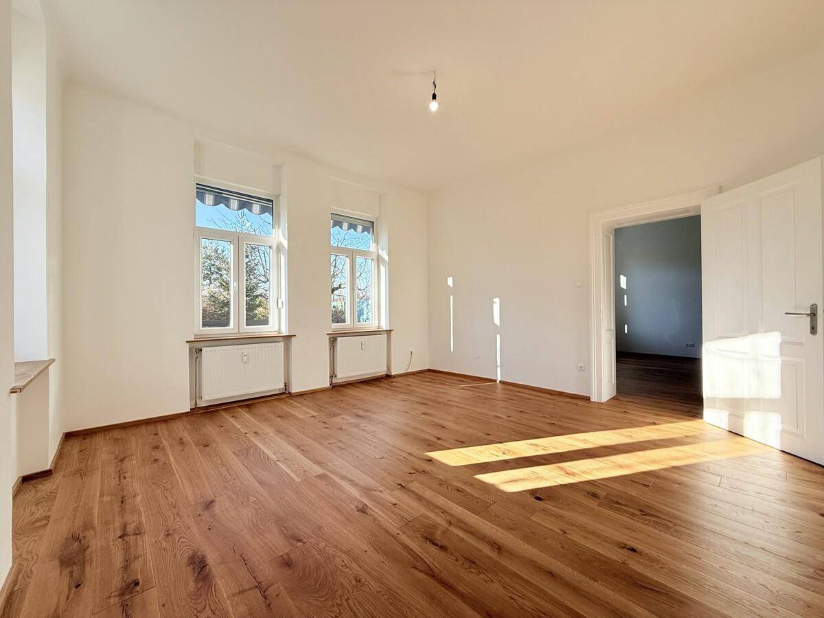 Geräumiges Wohnzimmer mit hellem Eichenparkett und zwei großen Fenstern, die den Raum lichtdurchfluten.