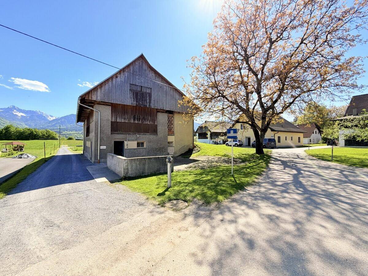 Rustikales Bauernhaus mit Holzbalkon und Steinfassade, umgeben von grünen Wiesen und Bergen im Hintergrund.