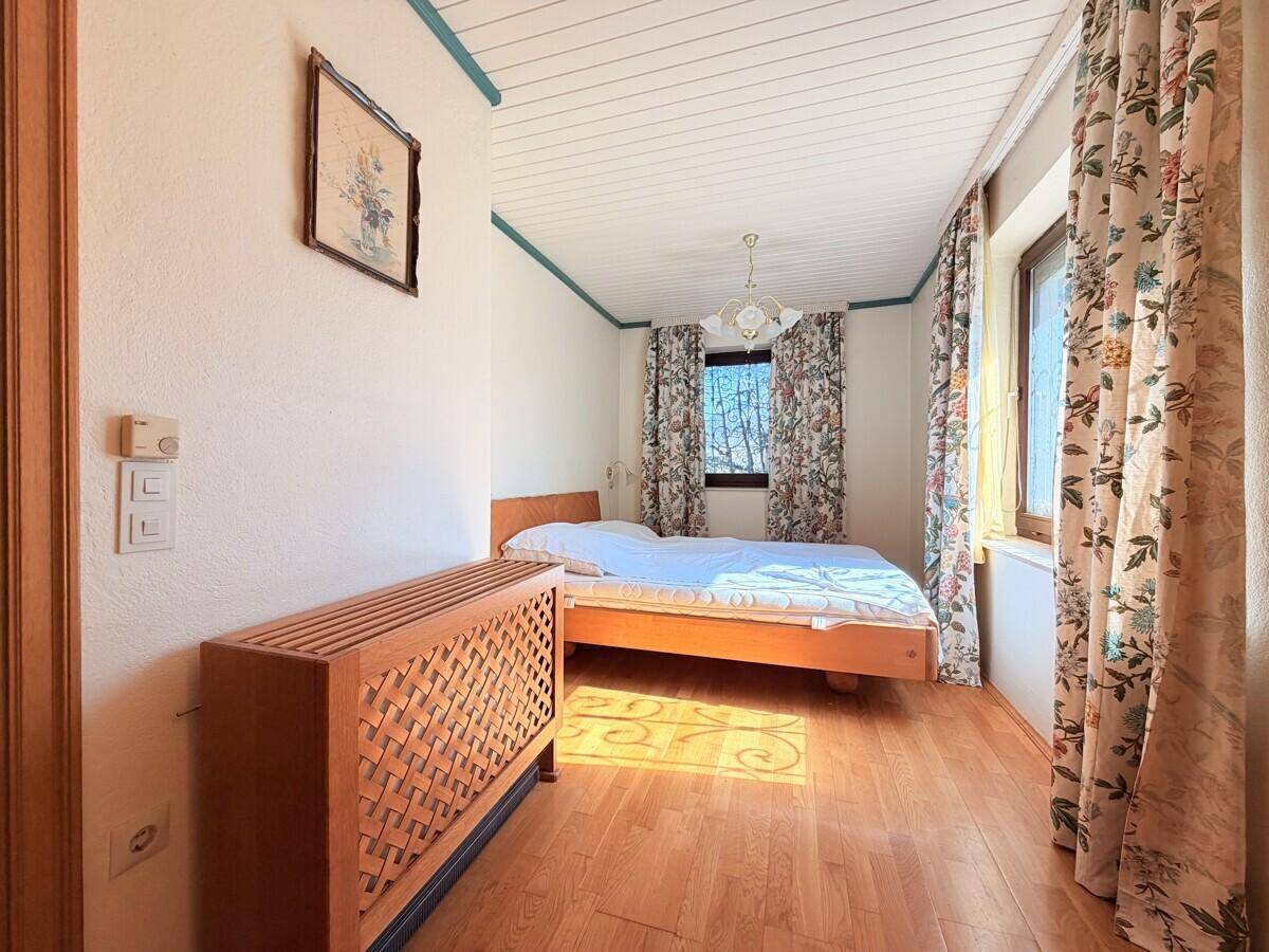 Schlafzimmer mit Holzboden, floralen Vorhängen und Blick ins Grüne durch das Fenster.