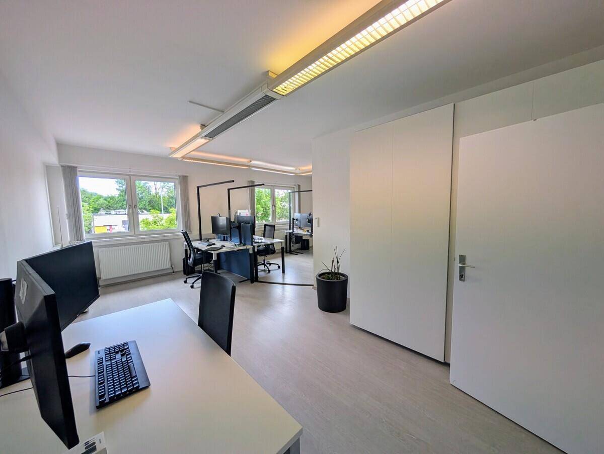 Helles, offenes Büro mit mehreren Arbeitsplätzen, großen Fenstern und integrierten Schränken.