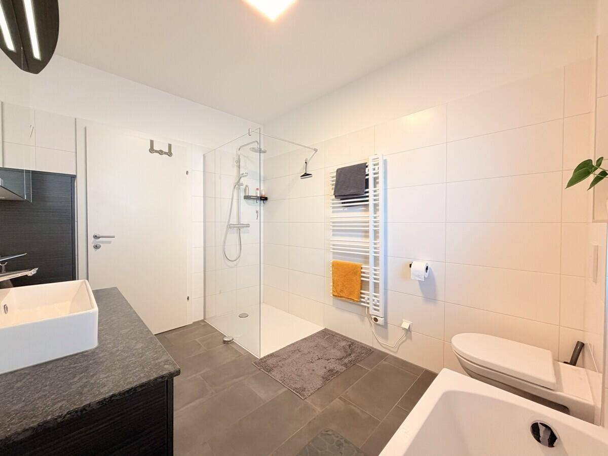 Geräumiges Badezimmer mit ebenerdiger Dusche, WC und modernem Waschtisch mit dunkler Arbeitsplatte.