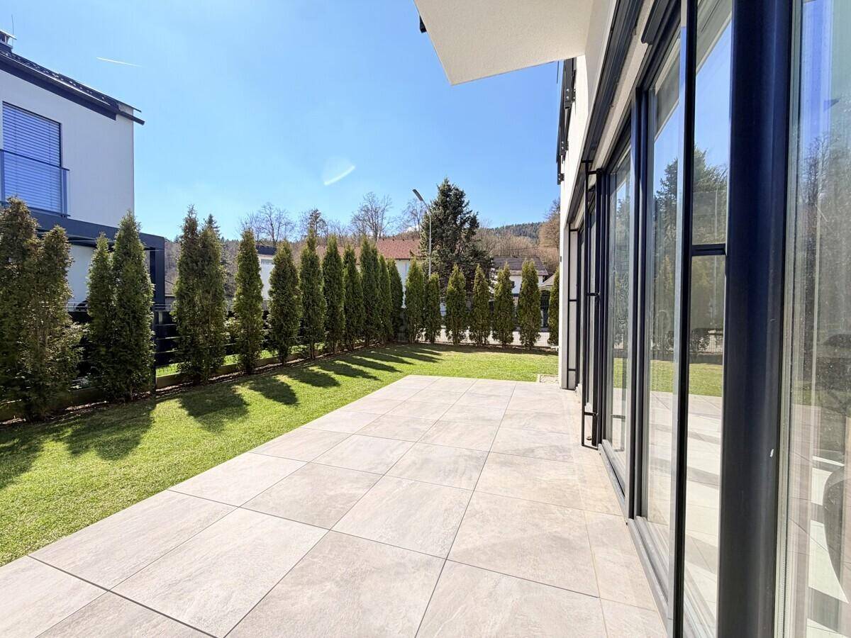 Sonniger Terrassenbereich mit großen Fliesen und angrenzendem Rasen, umgeben von Hecken.
