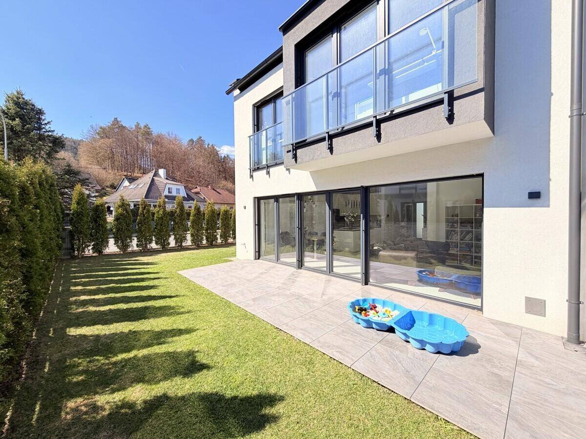 Sonniger Gartenbereich mit Rasen, großer Terrasse und Spielzeug für Kinder.