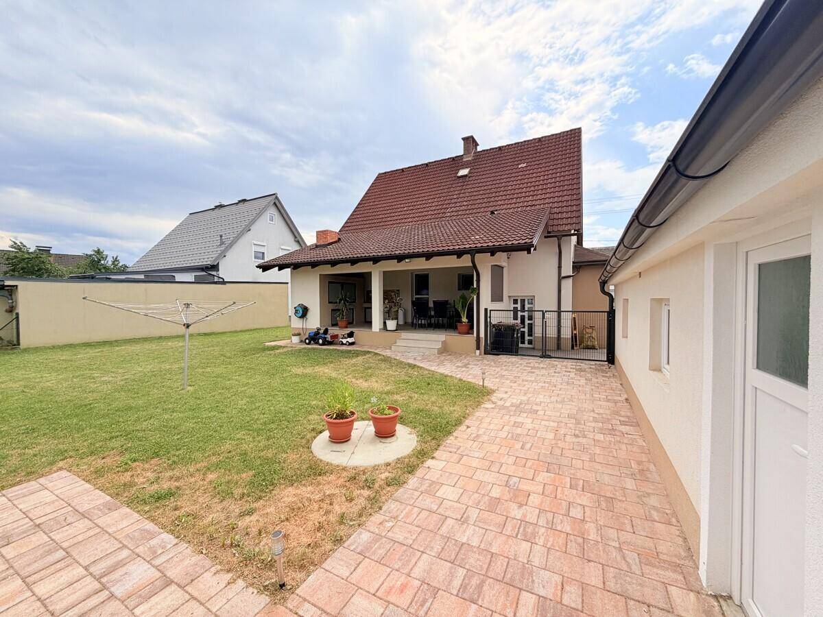 Gepflegter Gartenbereich mit Rasenfläche, gepflastertem Weg und überdachter Terrasse.