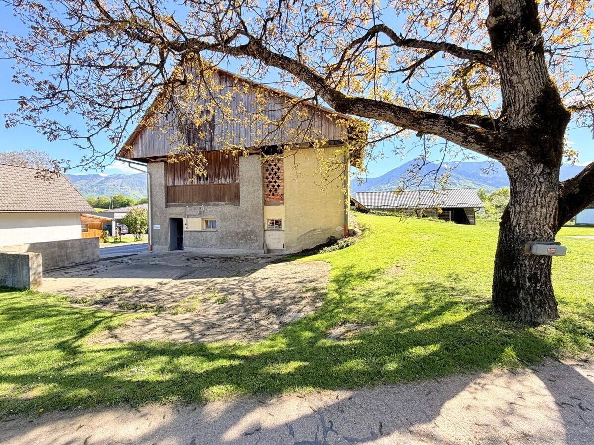 Altes Bauernhaus mit markantem Holzdach und großem Baum im Vordergrund, eingebettet in eine ländliche Umgebung.