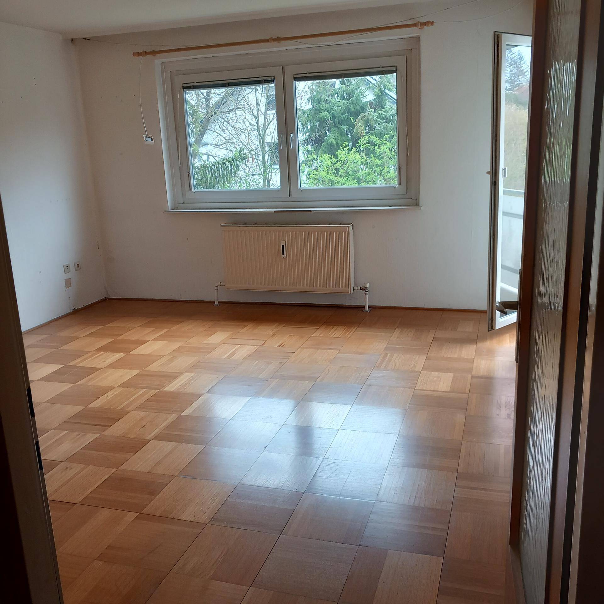 Heller, leerer Raum mit Parkettboden und großem Fenster, das viel Tageslicht hereinlässt.