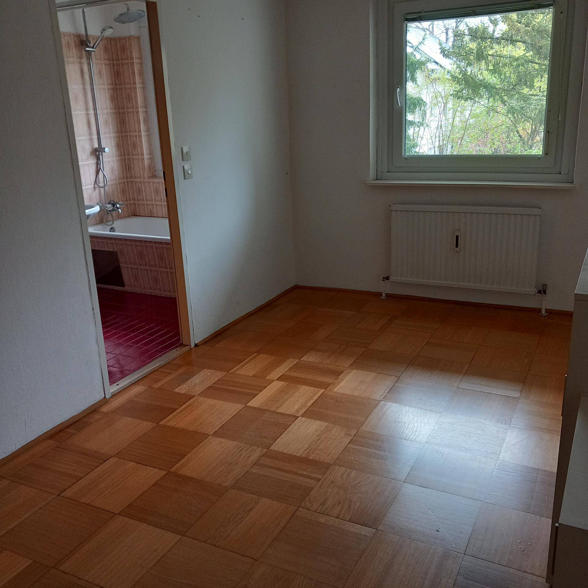 Leerer Raum mit Parkettboden und Fenster, angrenzend an ein Badezimmer mit roten Fliesen.