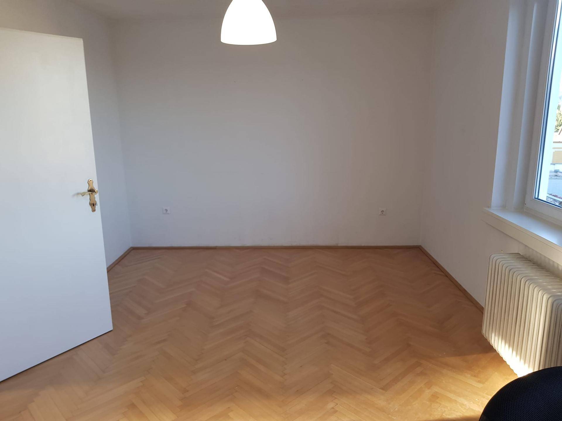 Helles, leeres Zimmer mit Fischgrätparkett, weißen Wänden und einem Fenster mit Heizkörper.