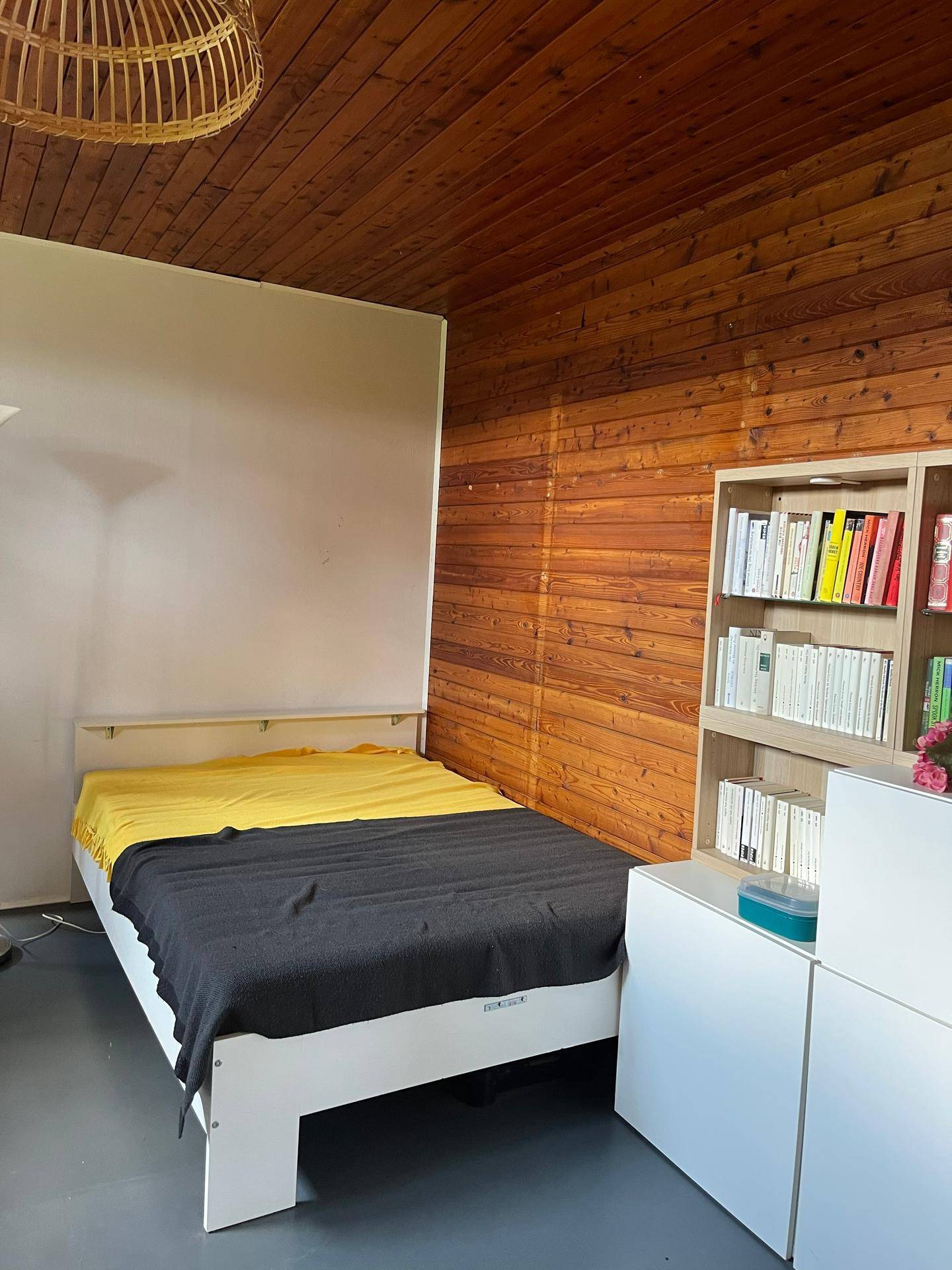 Schlafzimmer mit Holzwänden, Einzelbett und Bücherregal, beleuchtet durch ein Fenster.