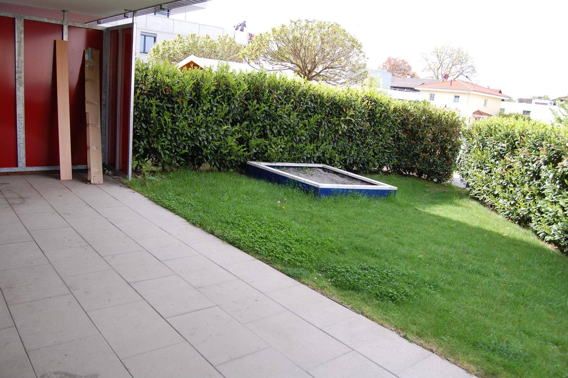Gartenbereich mit Sandkasten und Hecken, angrenzend an eine gepflasterte Terrasse.