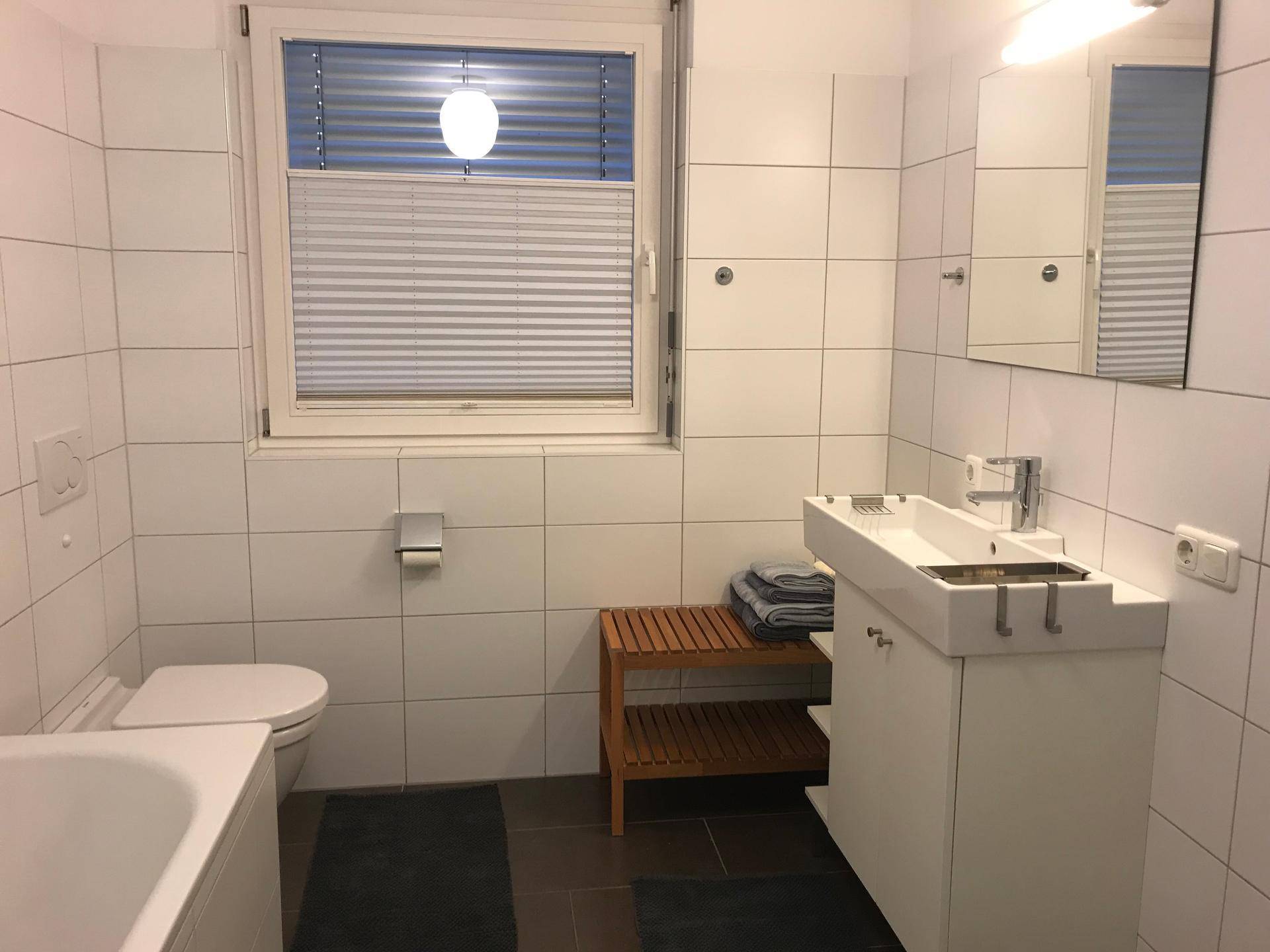 Helles Badezimmer mit weißen Fliesen, WC, Waschbecken und Fenster, ausgestattet mit Holzbank.