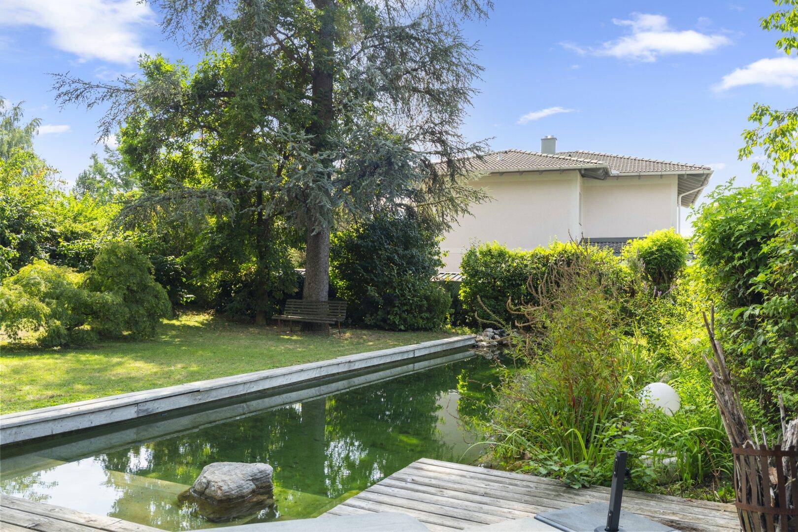 Weitläufiger Garten mit Teich, Rasenfläche und üppiger Bepflanzung unter blauem Himmel.