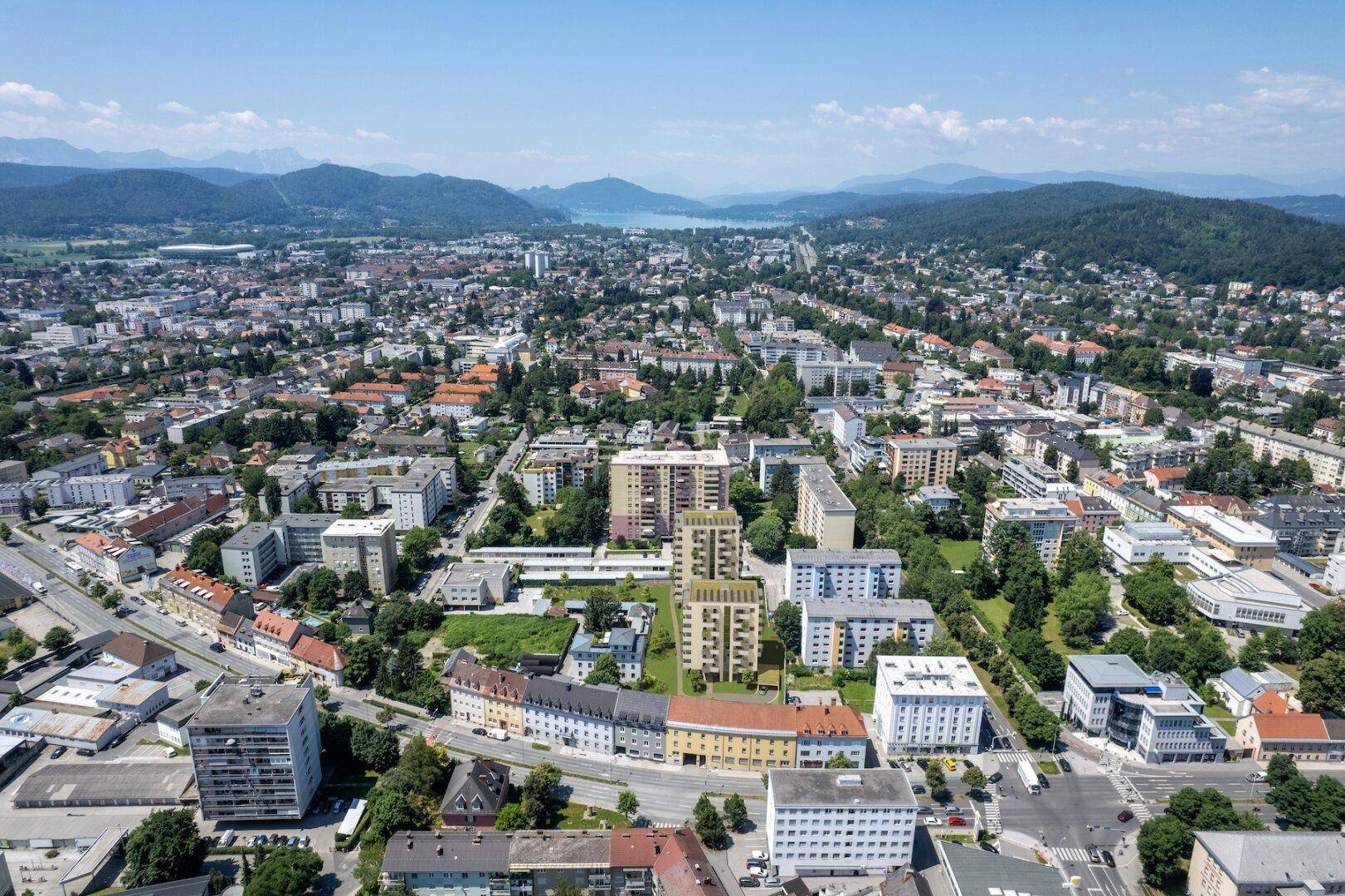 Weitläufige Luftaufnahme der Stadt mit den Wohngebäuden im Zentrum, umgeben von urbaner Bebauung und einem See in der Ferne.