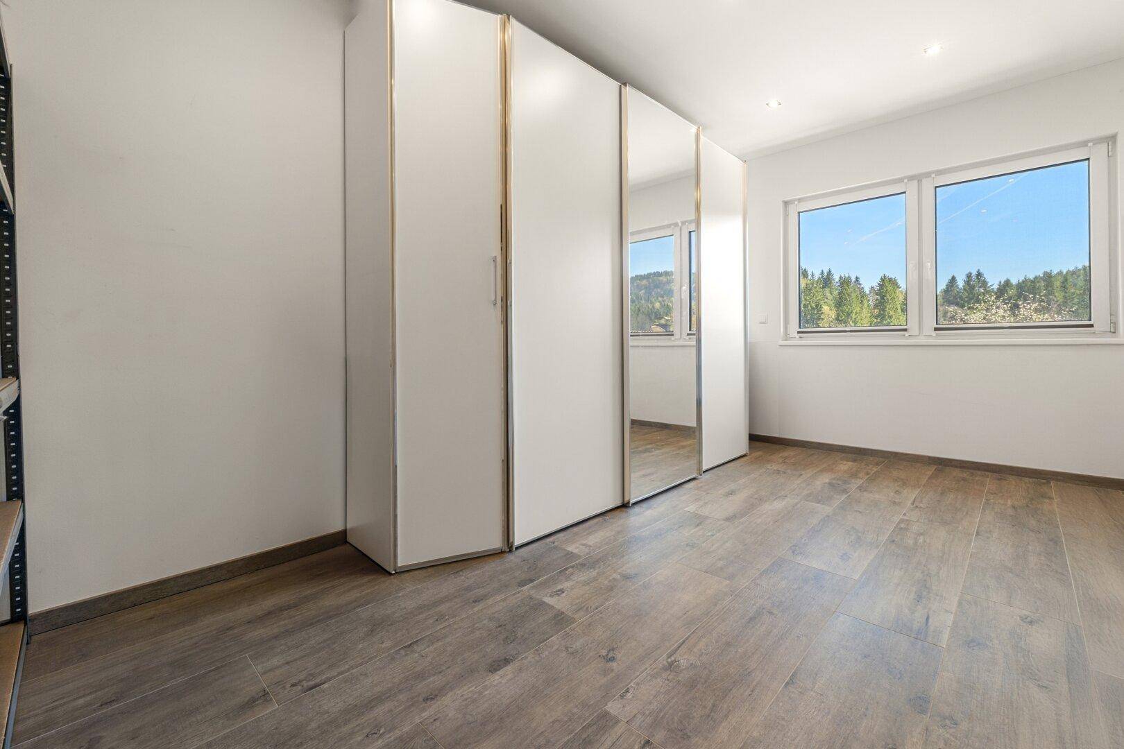 Geräumiges Zimmer mit Einbauschränken, Holzoptik-Fliesenboden und großen Fenstern.