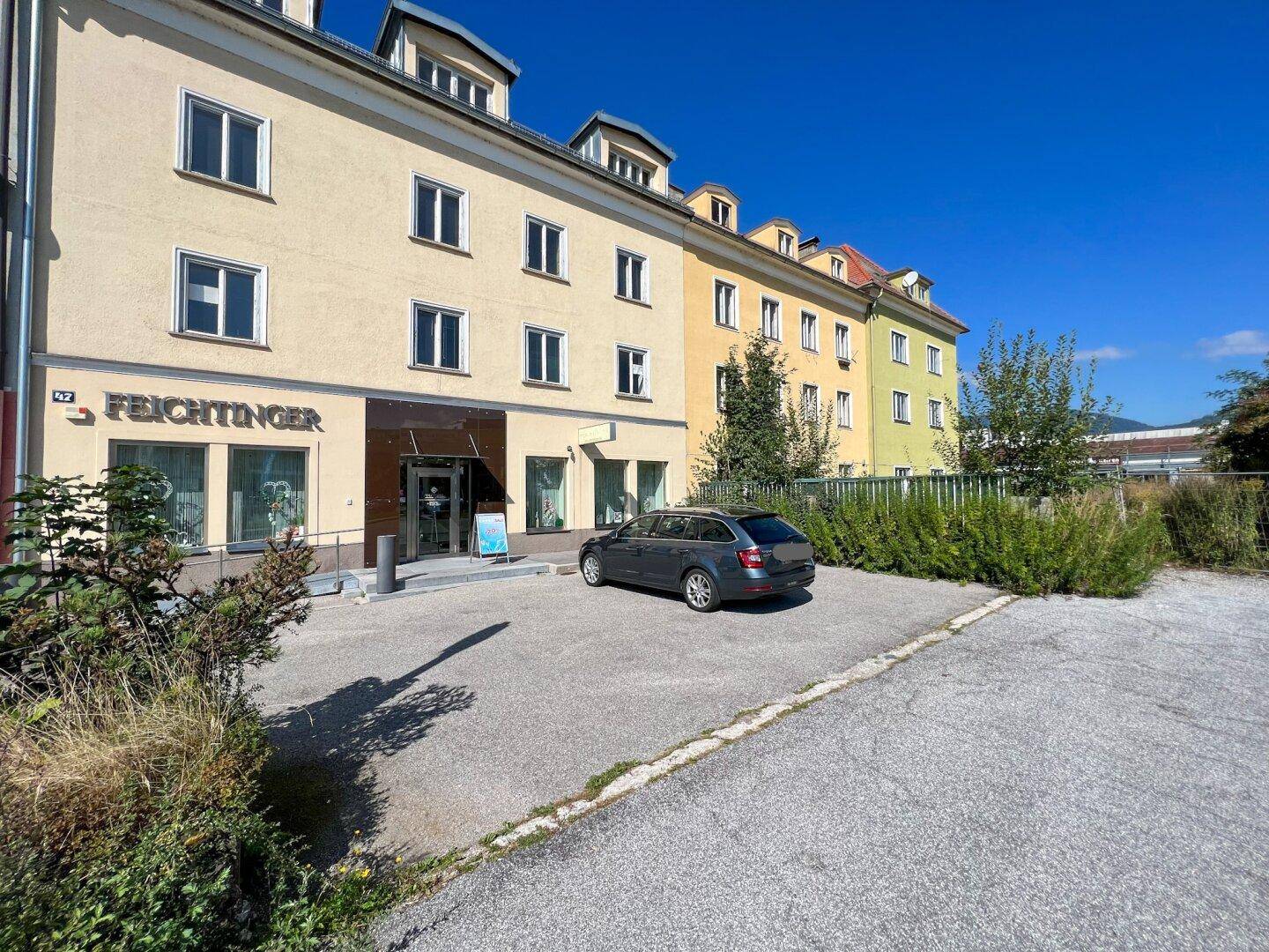 Ein mehrstöckiges Gebäude mit Geschäften im Erdgeschoss und Wohnungen darüber, umgeben von einem Parkplatz.