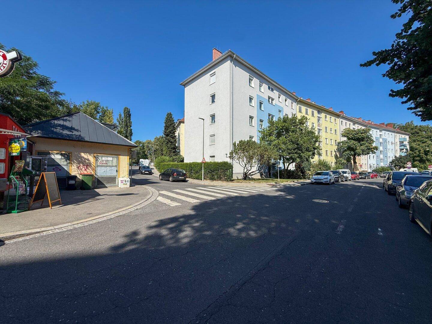 Blick auf eine Wohnstraße mit Gebäuden und einem kleinen Kiosk unter blauem Himmel.