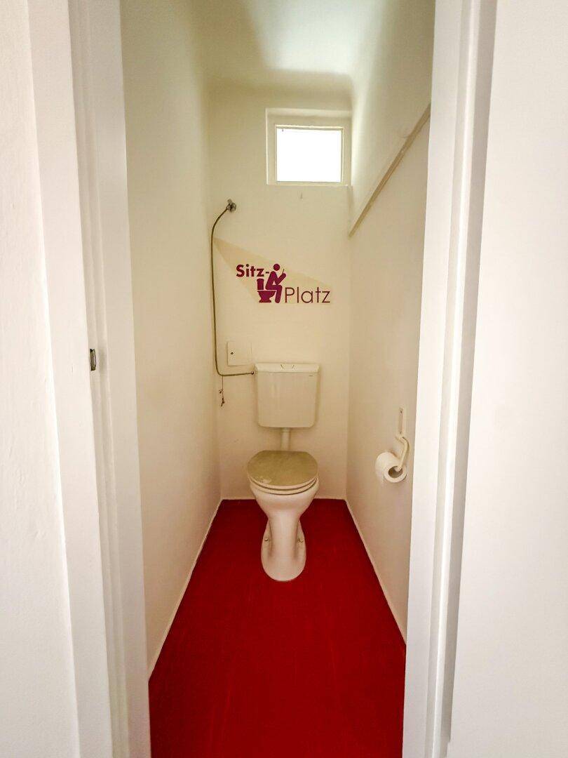 Kleines separates WC mit rotem Boden und einem Fenster für natürliche Belüftung.