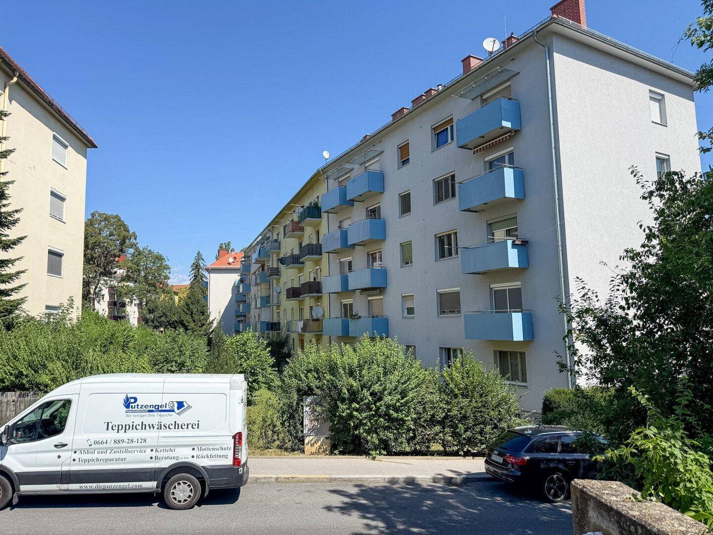 Mehrfamilienhaus mit blauen Balkonen und grüner Bepflanzung an einem sonnigen Tag.