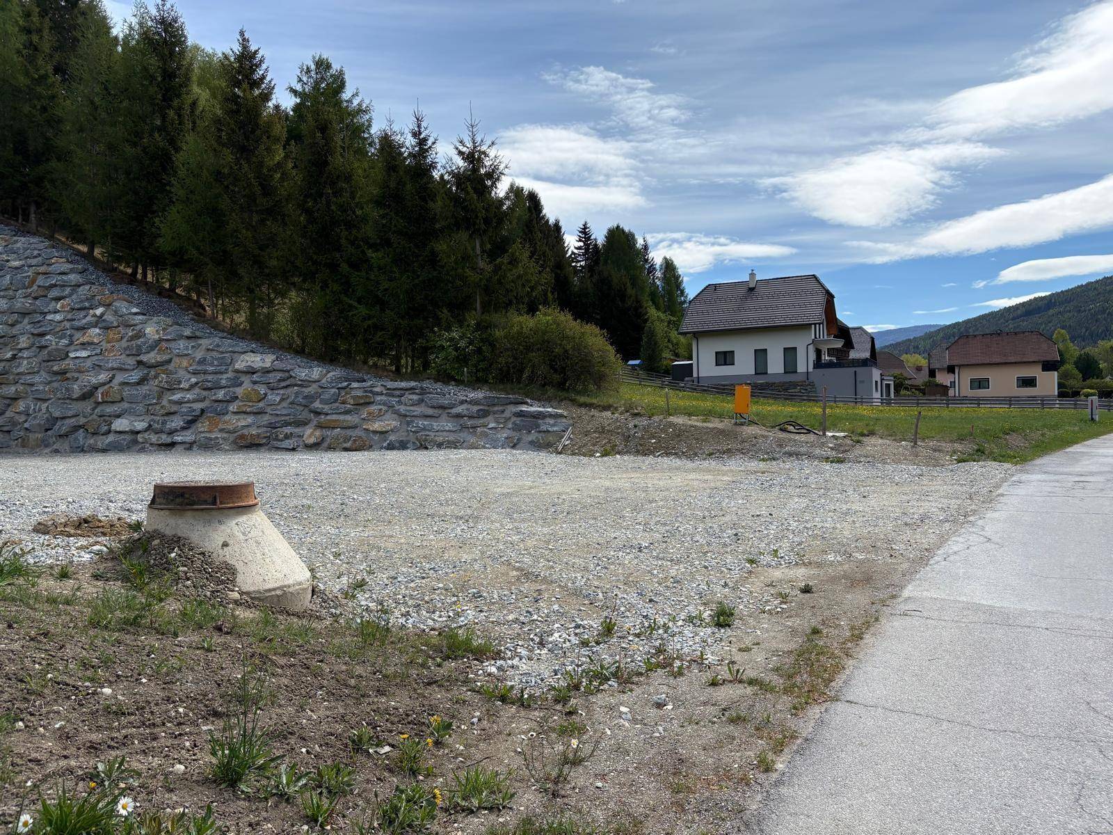 Schotterfläche mit einem Betonschacht und einer Natursteinmauer, im Hintergrund ein Wohnhaus.