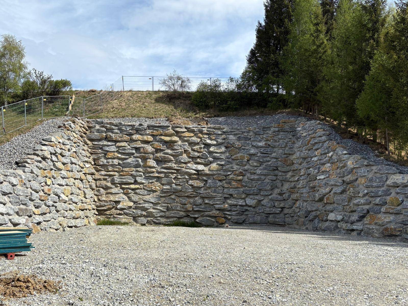 Eine massive Natursteinmauer begrenzt den Bereich, mit Schotterfläche davor und Bäumen im Hintergrund.
