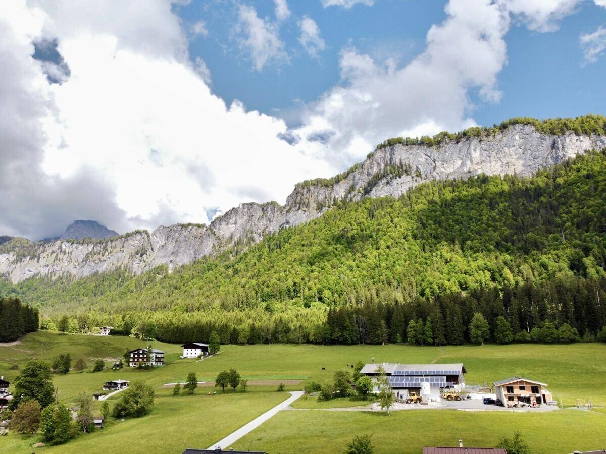 Weitläufige Landschaft mit grünen Hügeln, bewaldeten Bergen und vereinzelten Häusern unter blauem Himmel.