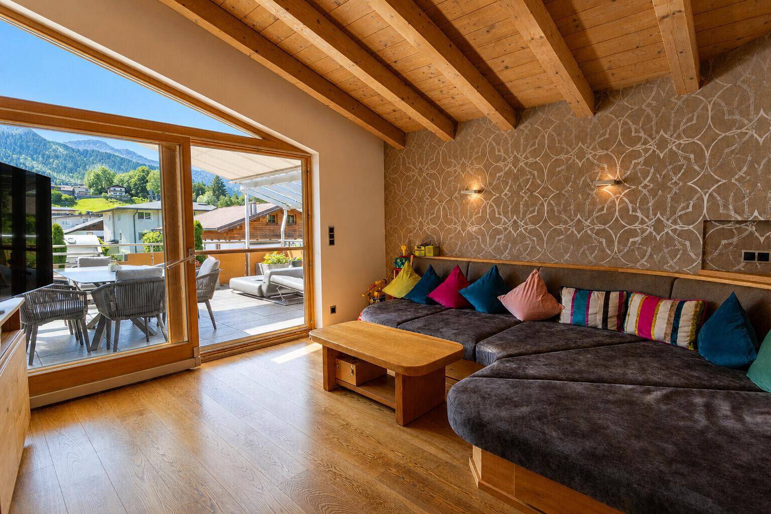 Gemütliches Wohnzimmer mit Holzbalken, großem Sofa und Zugang zu einer sonnigen Terrasse mit Bergblick.