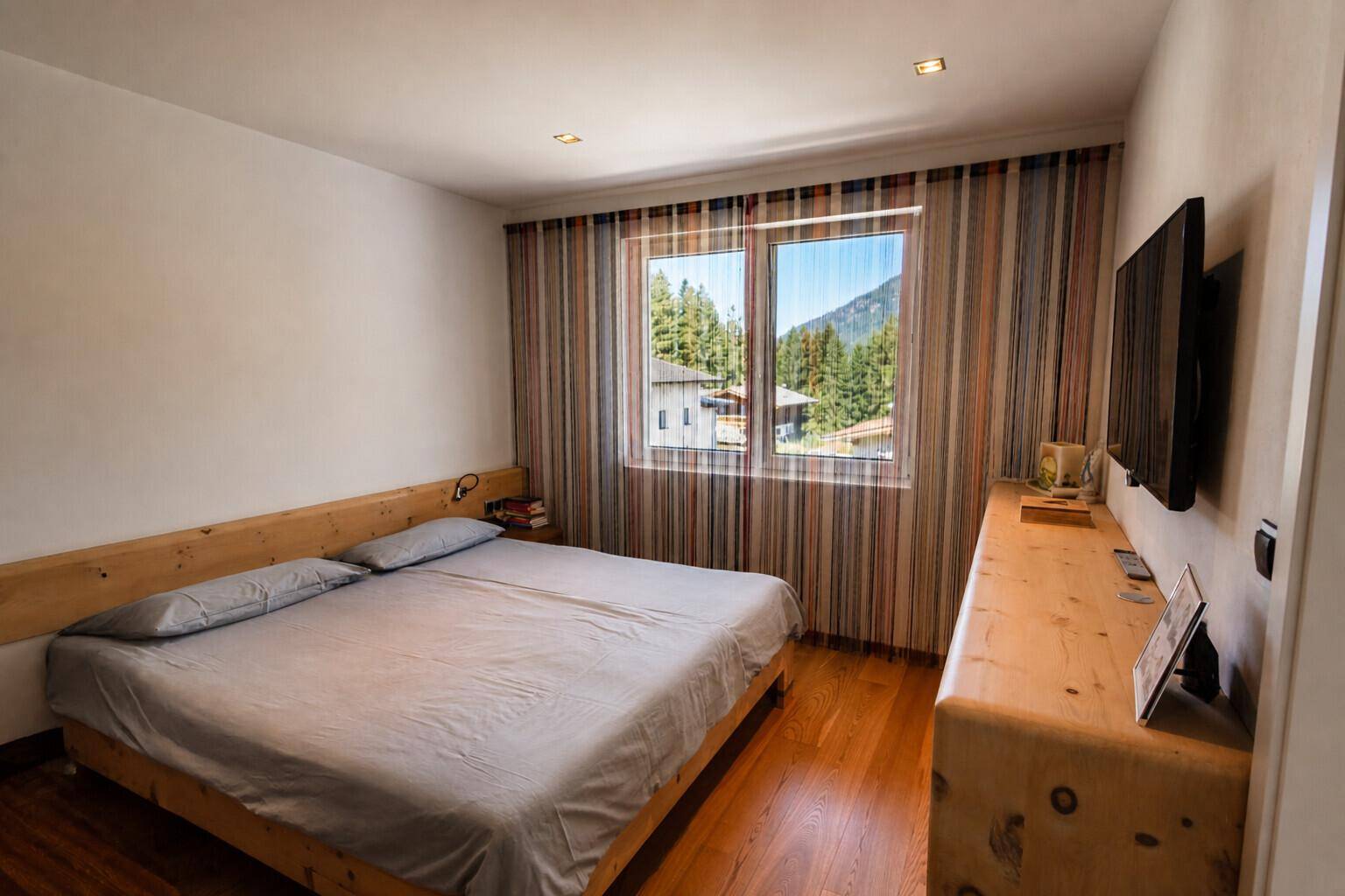 Helles Schlafzimmer mit Holzbett, Holzkommode und Blick auf die bewaldeten Berge.