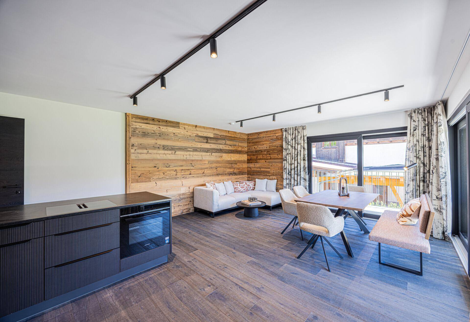 Heller Wohn- und Essbereich mit Holzwand, moderner Küchenzeile und Zugang zum Balkon.