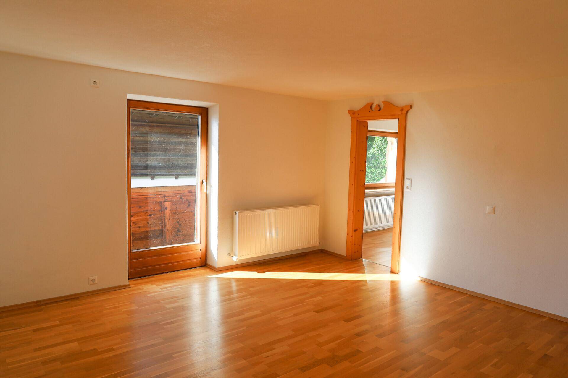 Heller, leerer Raum mit Parkettboden und Zugang zu einem Balkon, durchflutet von natürlichem Sonnenlicht.