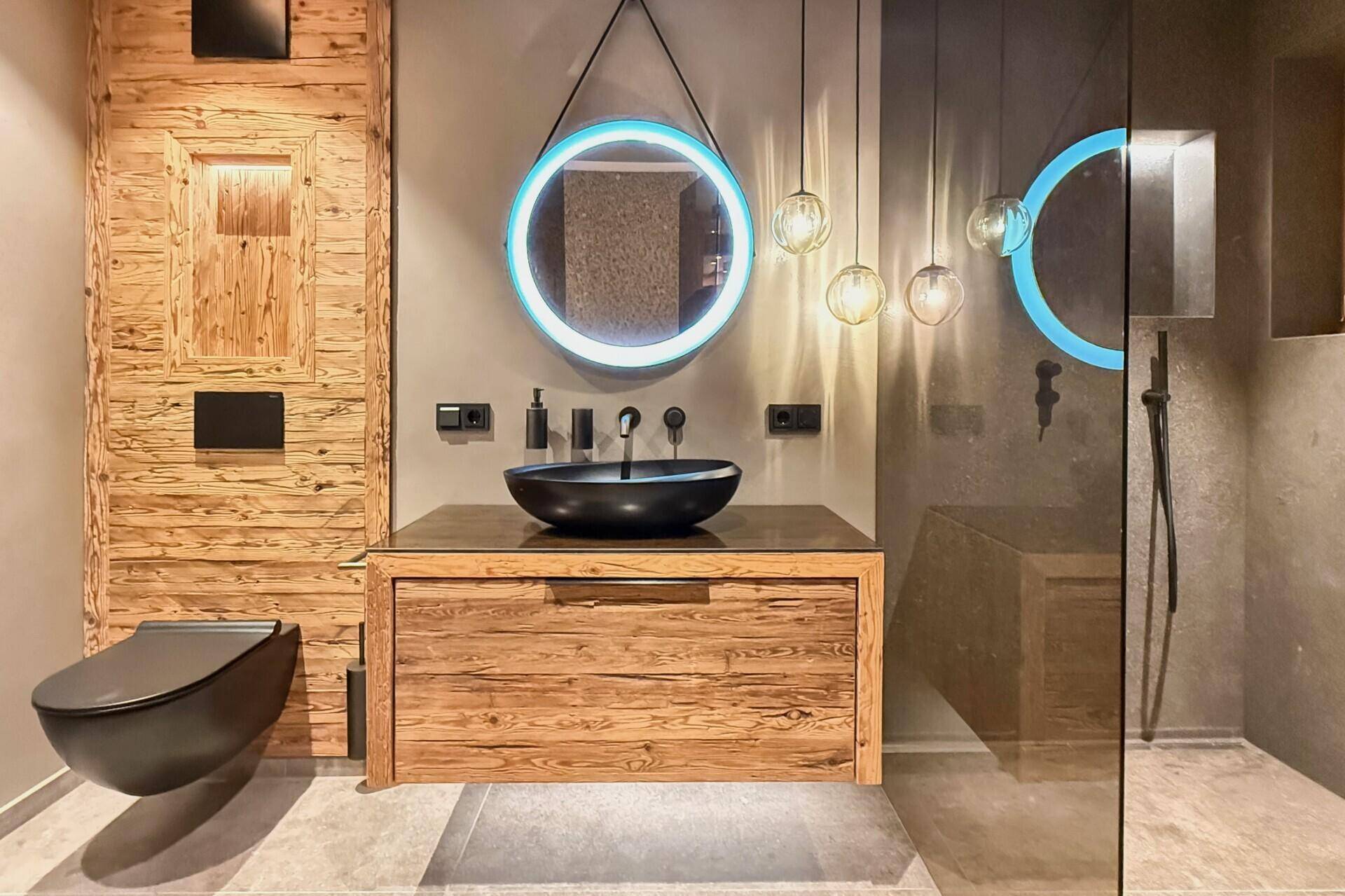 Modernes Badezimmer mit beleuchtetem Spiegel, schwarzem Waschbecken und Holzelementen.