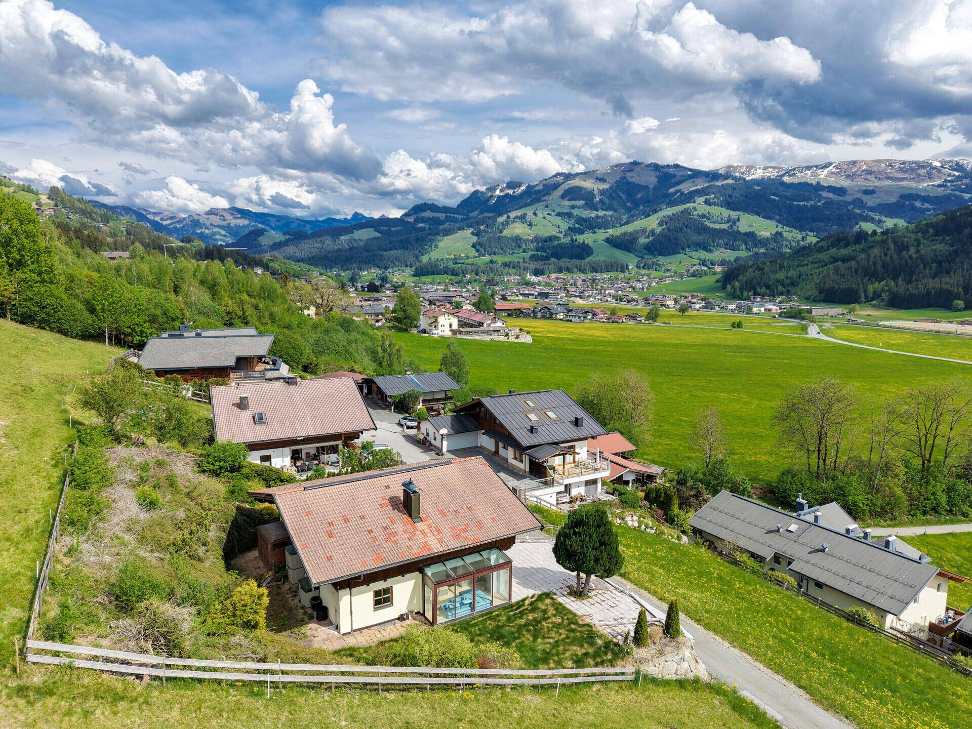Panoramablick auf eine Wohnsiedlung am Hang mit grünen Wiesen und einem Dorf vor alpiner Kulisse.