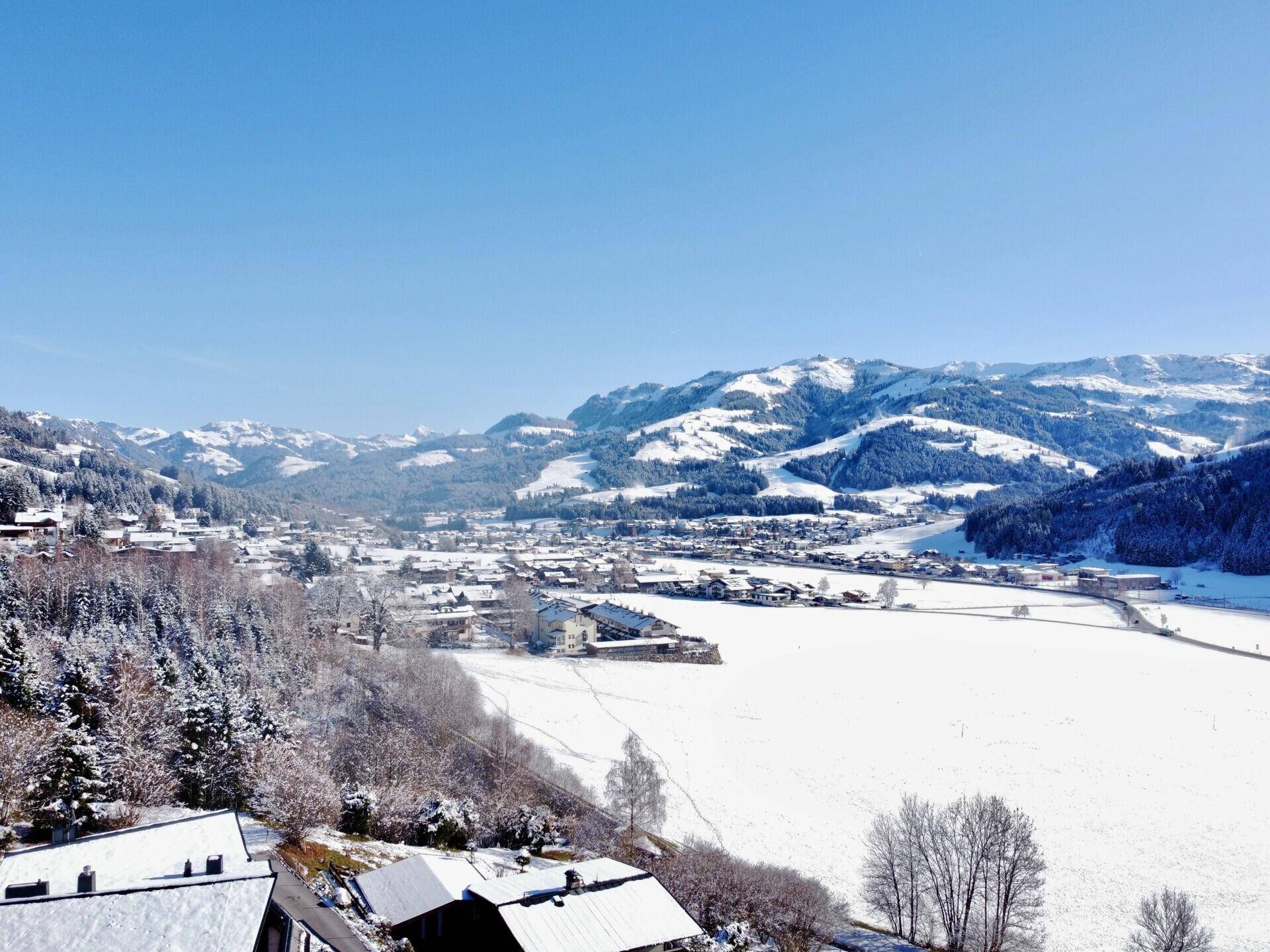 Panoramablick auf eine verschneite Landschaft mit einem Dorf im Tal, umgeben von bewaldeten Bergen.