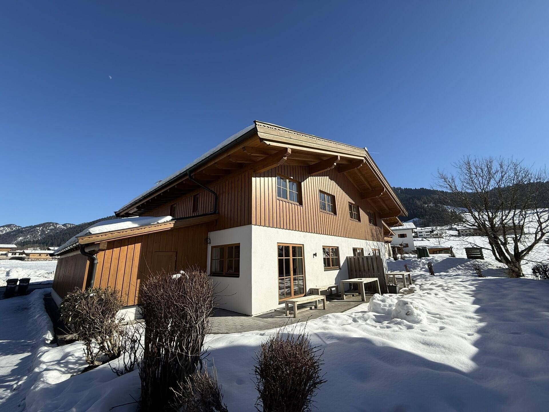 Seitenansicht des Chalets mit kleiner Terrasse und verschneitem Gartenbereich an einem sonnigen Tag.
