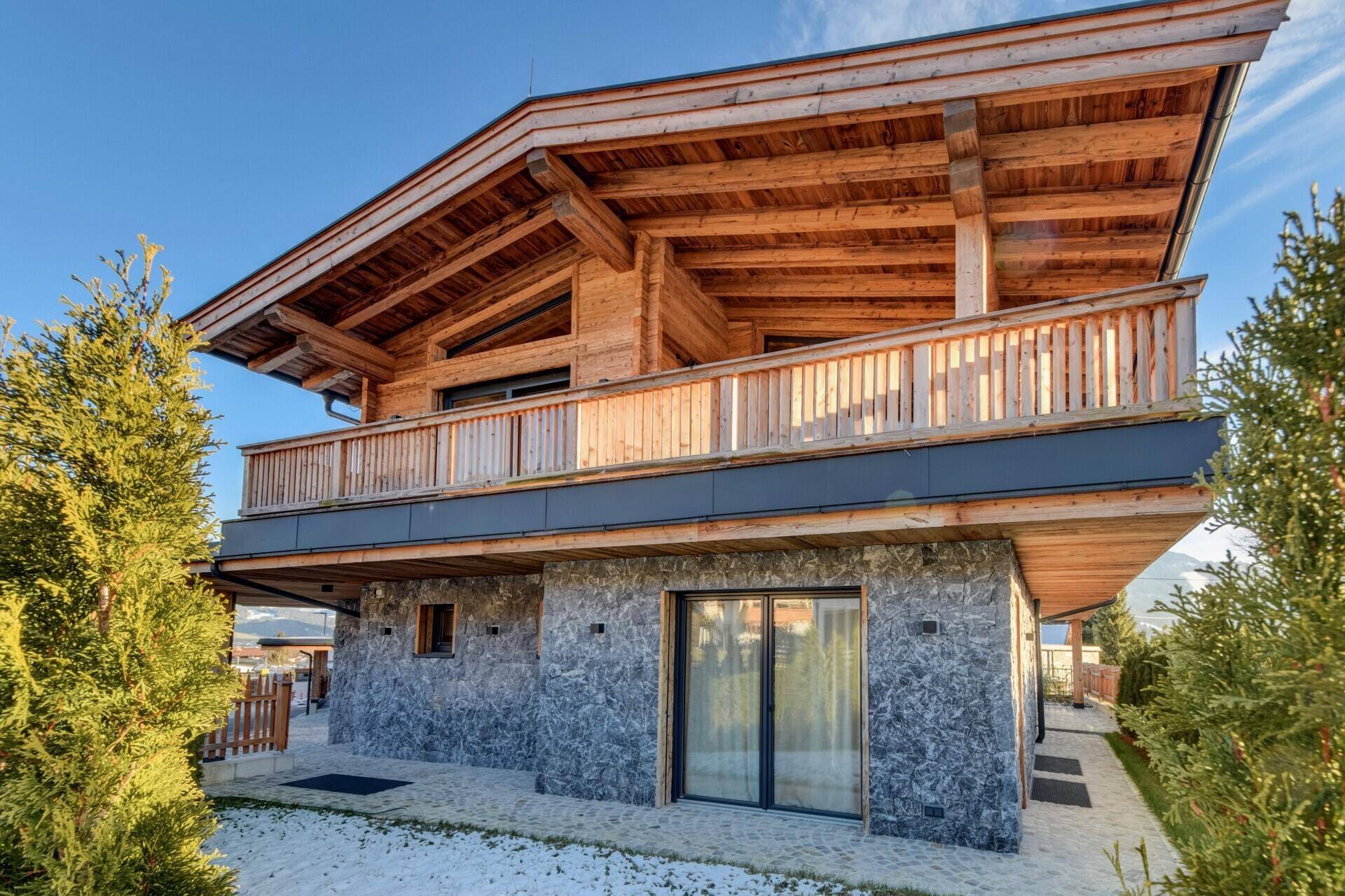 Modernes Chalet mit Holz- und Steinfassade in sonniger Lage, umgeben von grünen Pflanzen.