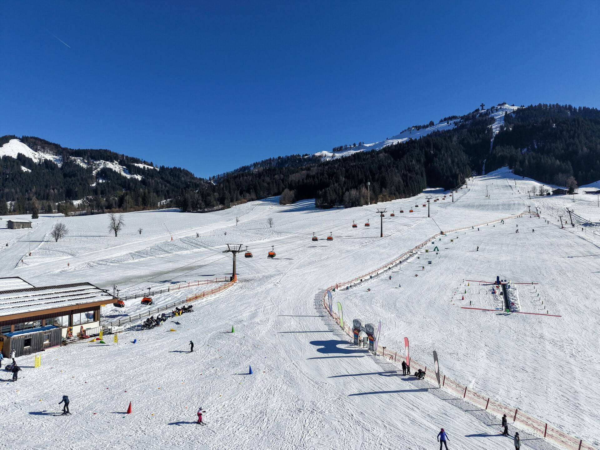 Weitläufige Skipiste mit Skiliften und Skifahrern unter blauem Himmel an einem sonnigen Wintertag.
