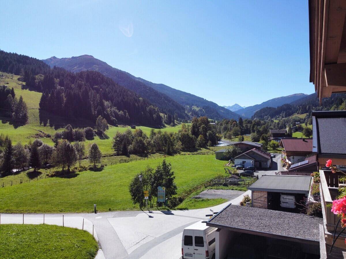 Panoramablick über ein grünes Tal mit bewaldeten Bergen und vereinzelten Häusern unter blauem Himmel.