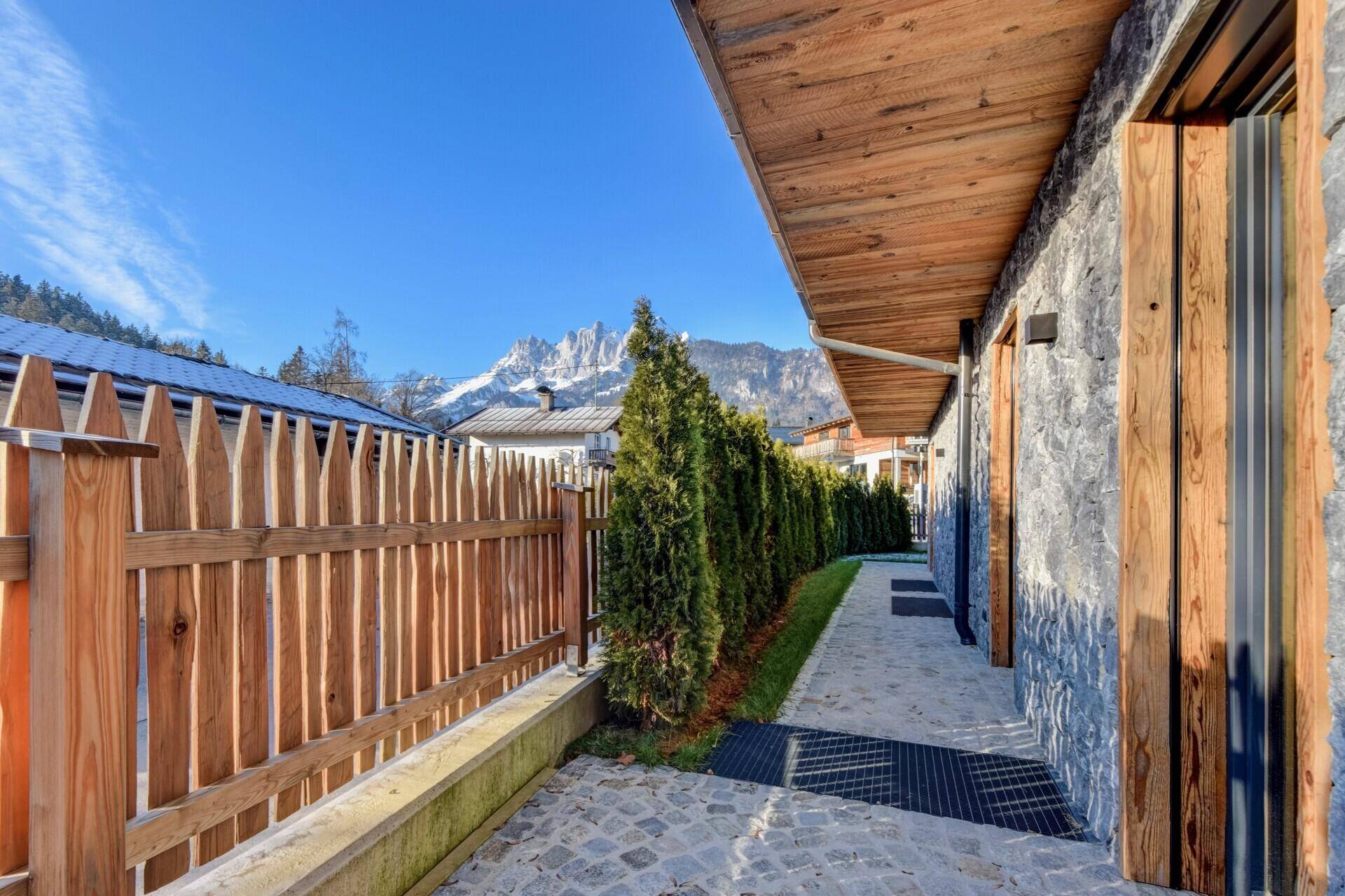 Seitlicher Zugang zum Haus mit Steinweg und Holzzaun, umgeben von Bäumen und Hecken.