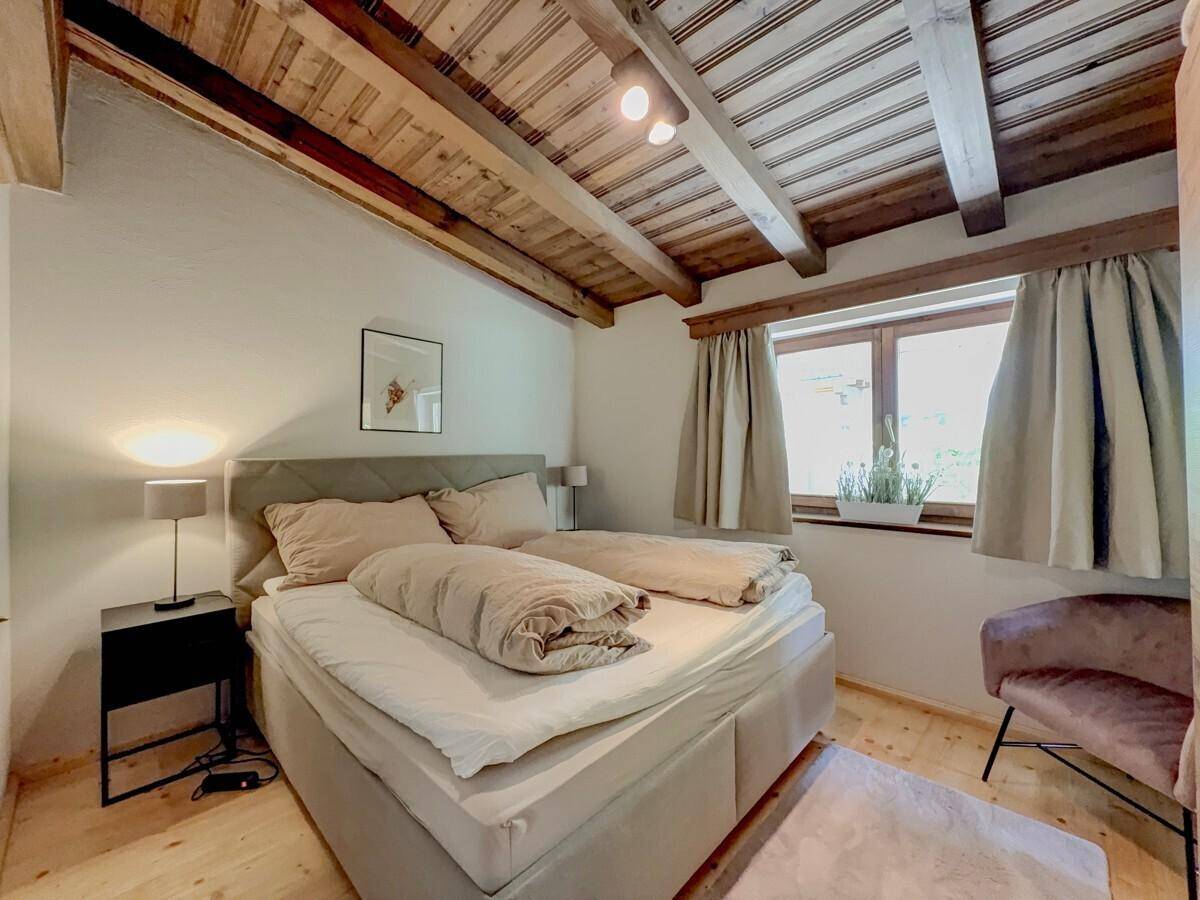 Schlafzimmer mit Holzbalkendecke, Doppelbett und einem Fenster mit Blick ins Grüne.