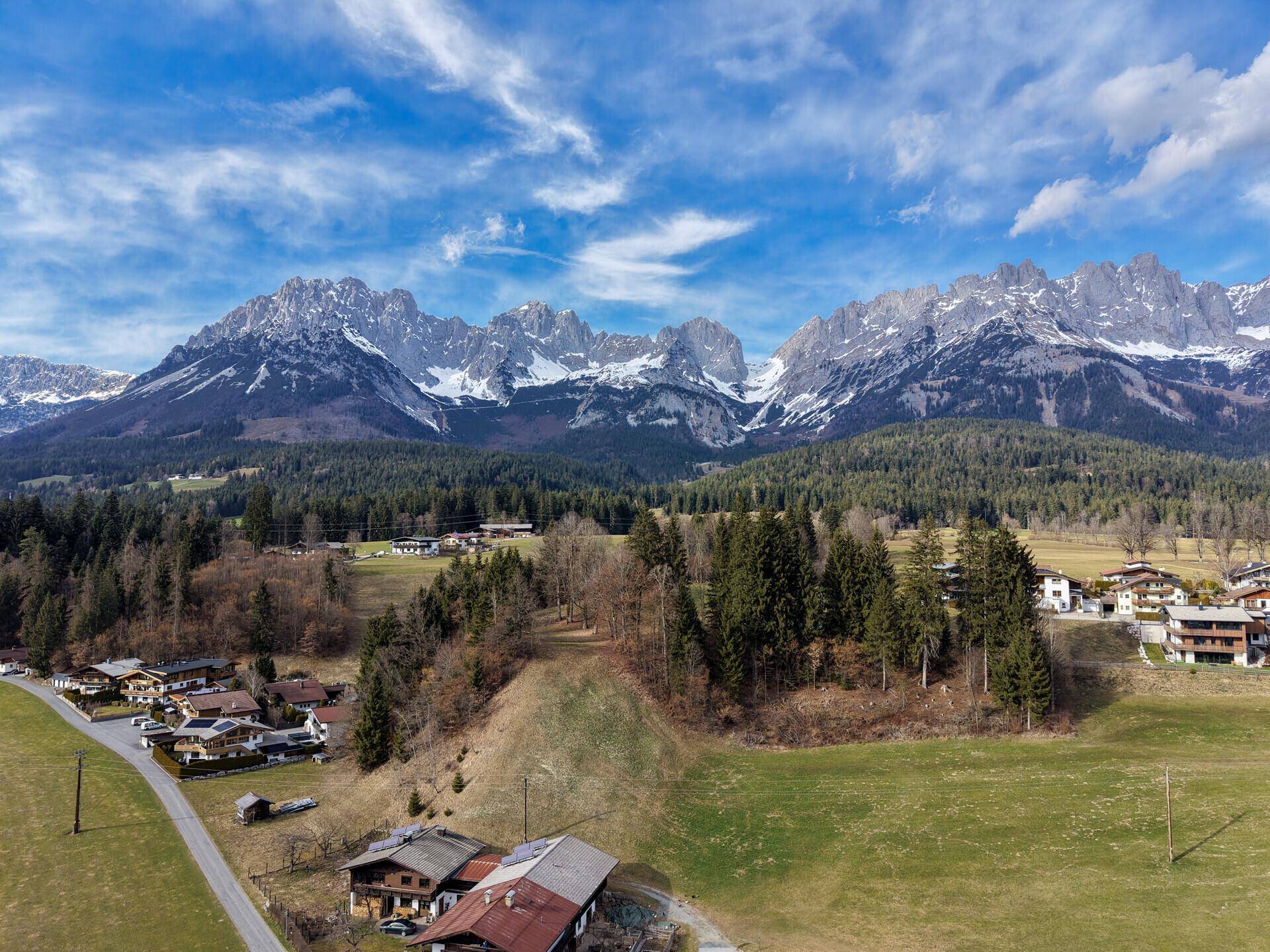 Weitläufige Landschaft mit traditionellen Häusern und majestätischen Bergen unter blauem Himmel.