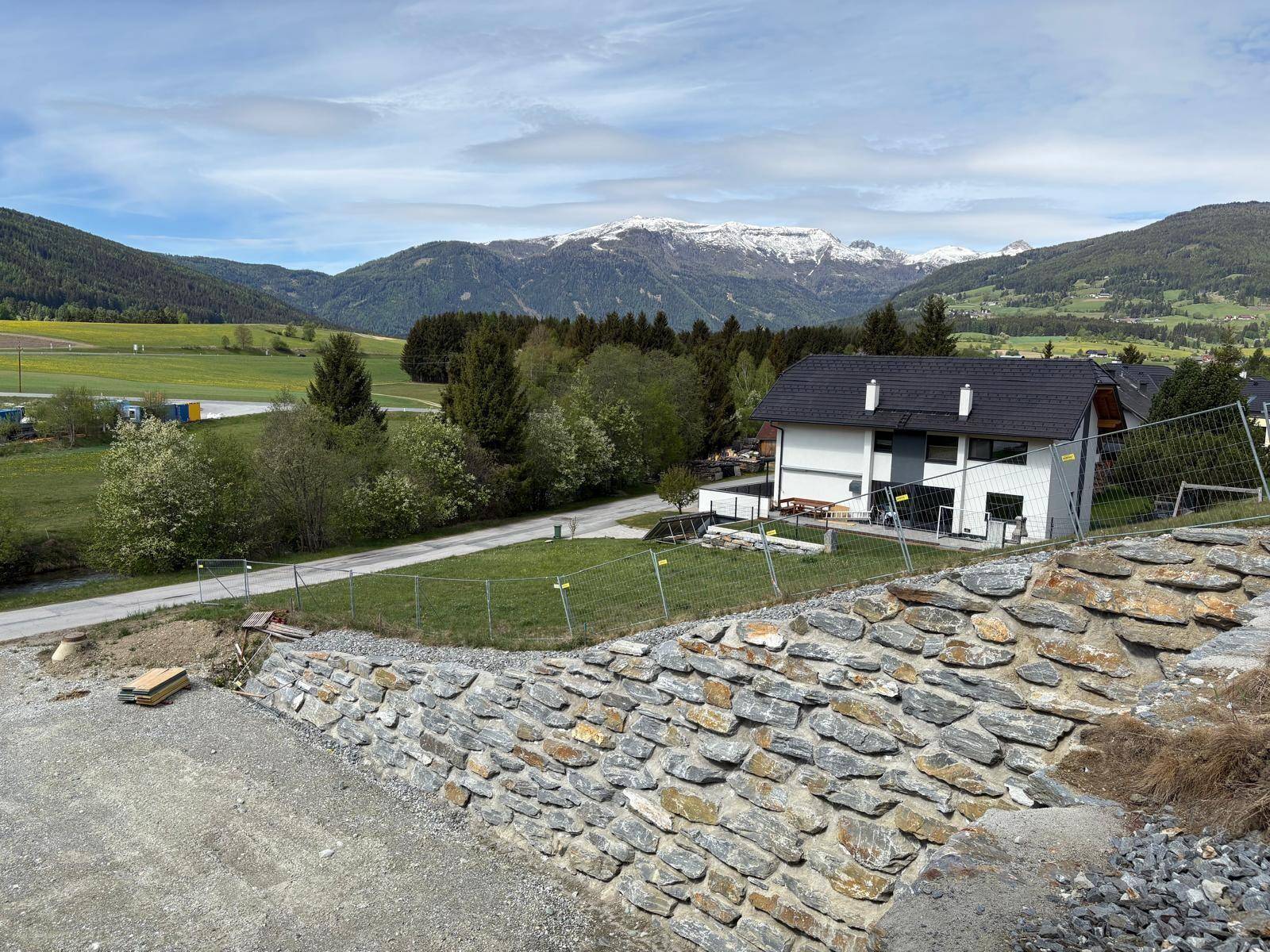Modernes Haus mit schwarzem Dach und weißer Fassade, umgeben von grüner Landschaft und Bergen.