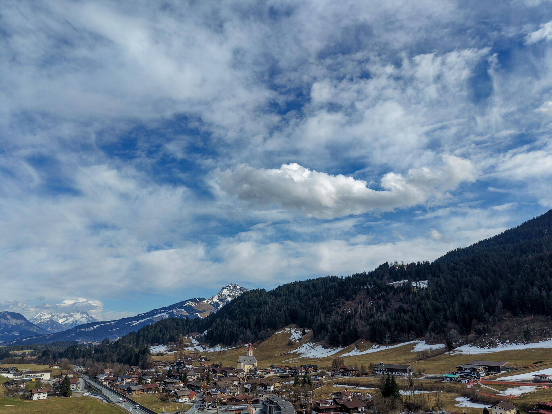 Blick über eine kleine Stadt im Tal mit schneebedeckten Bergen und bewaldeten Hängen im Hintergrund.