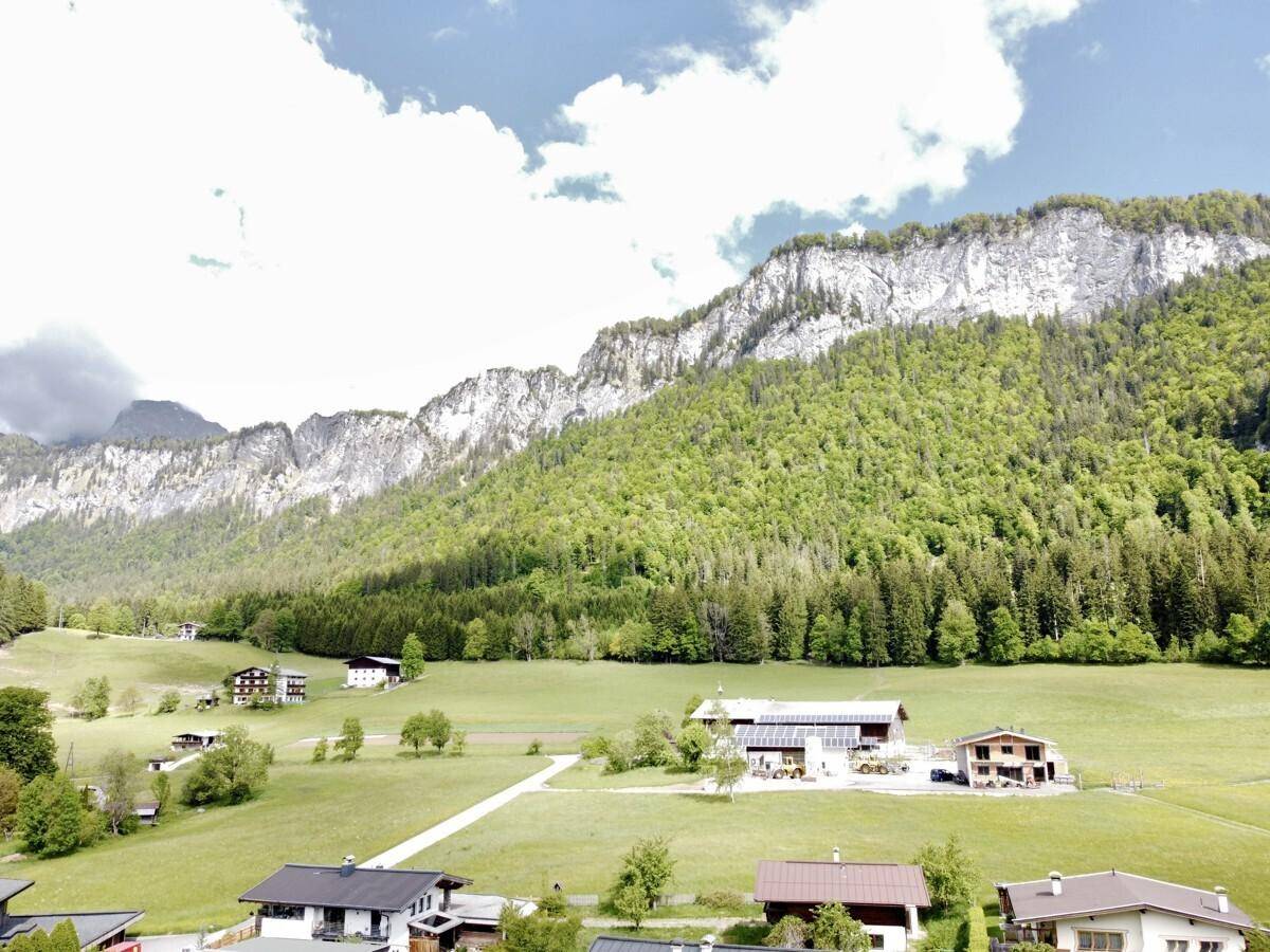 Weitläufige Landschaft mit grünen Hügeln, bewaldeten Bergen und vereinzelten Häusern unter blauem Himmel.