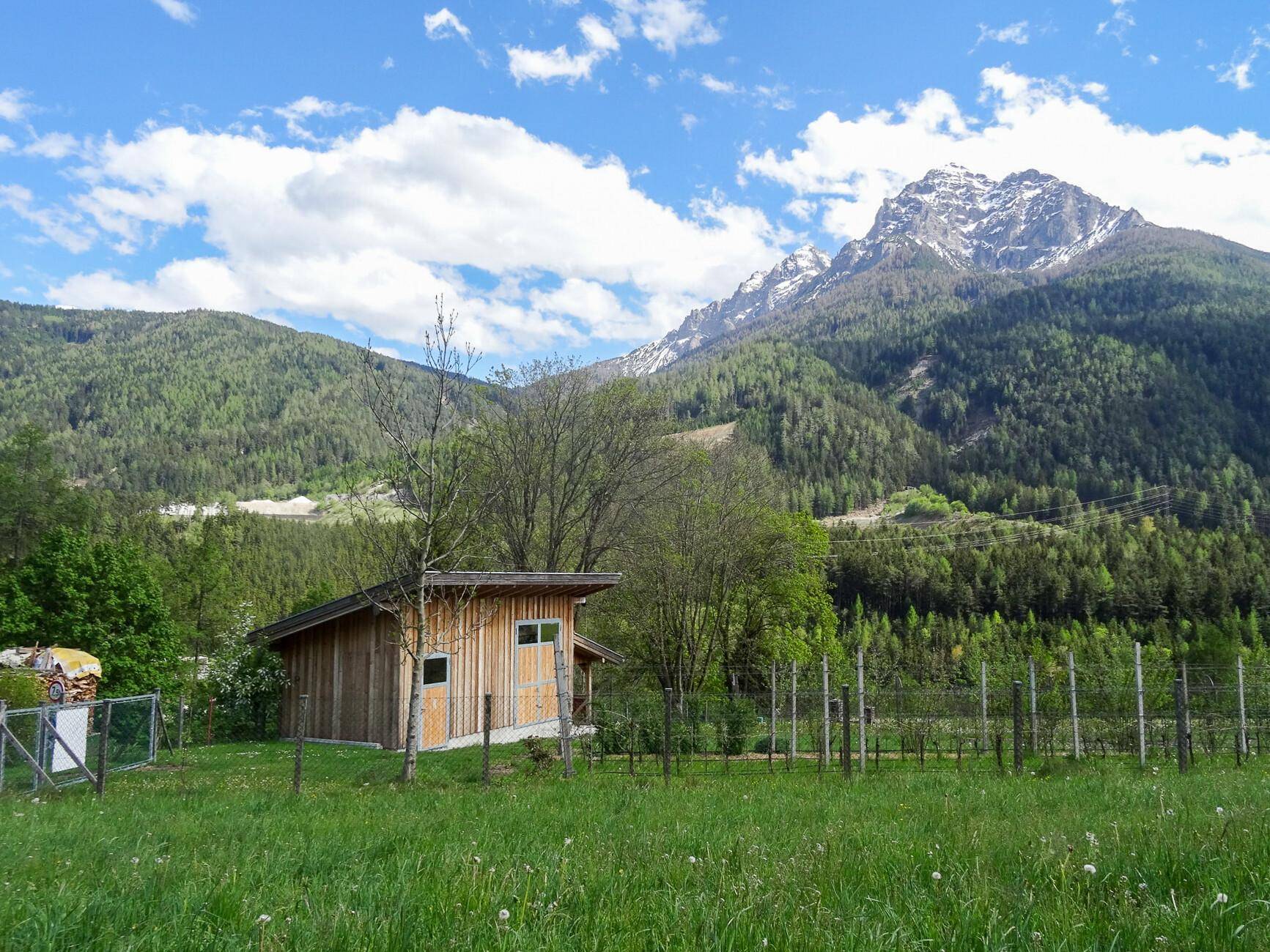 Kleiner Holzschuppen auf einer grünen Wiese mit beeindruckender Bergkulisse und Wald.