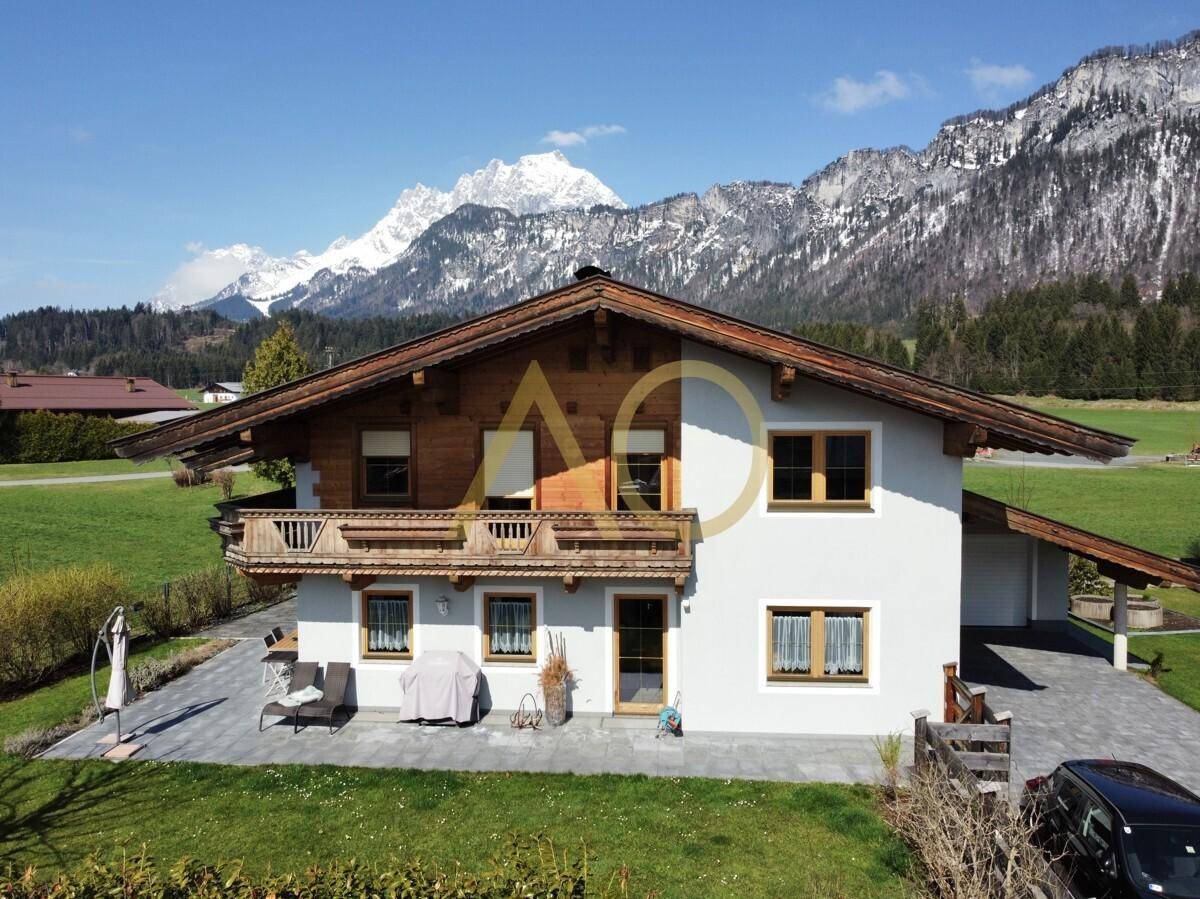 Traditionelles Haus mit Holzbalkon und gepflasterter Terrasse vor majestätischer Bergkulisse.