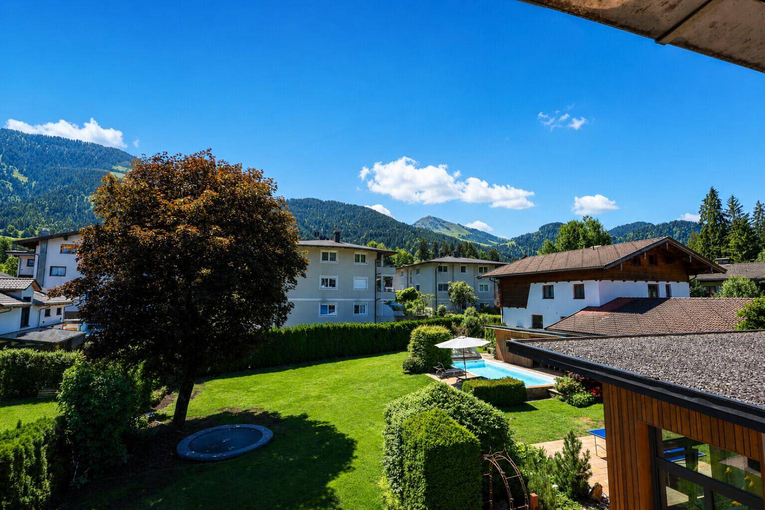 Großer gepflegter Garten mit Pool, Trampolin und Bergblick, umgeben von grünen Hecken und Bäumen.