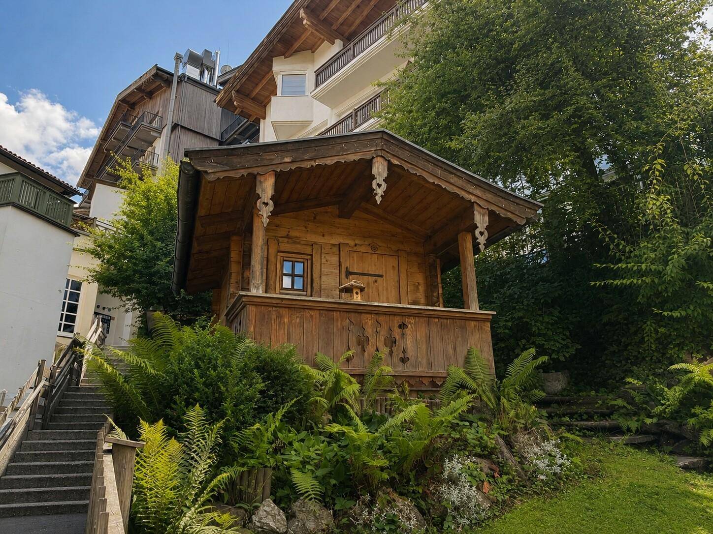 Rustikaler Holzpavillon mit geschnitzten Verzierungen, eingebettet in eine grüne Hanglandschaft mit Treppenaufgang.