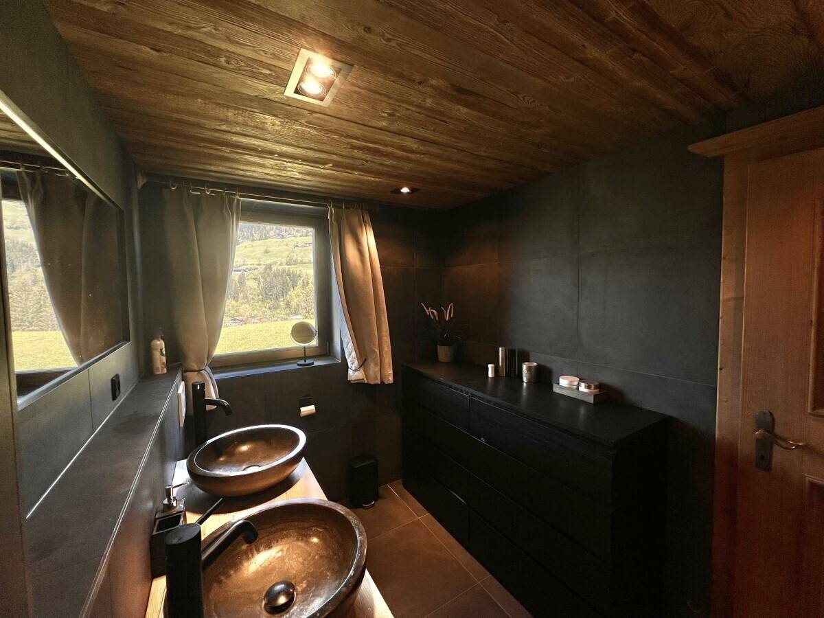 Modernes Badezimmer mit dunklen Fliesen, Holzbalkendecke und zwei Aufsatzwaschbecken.