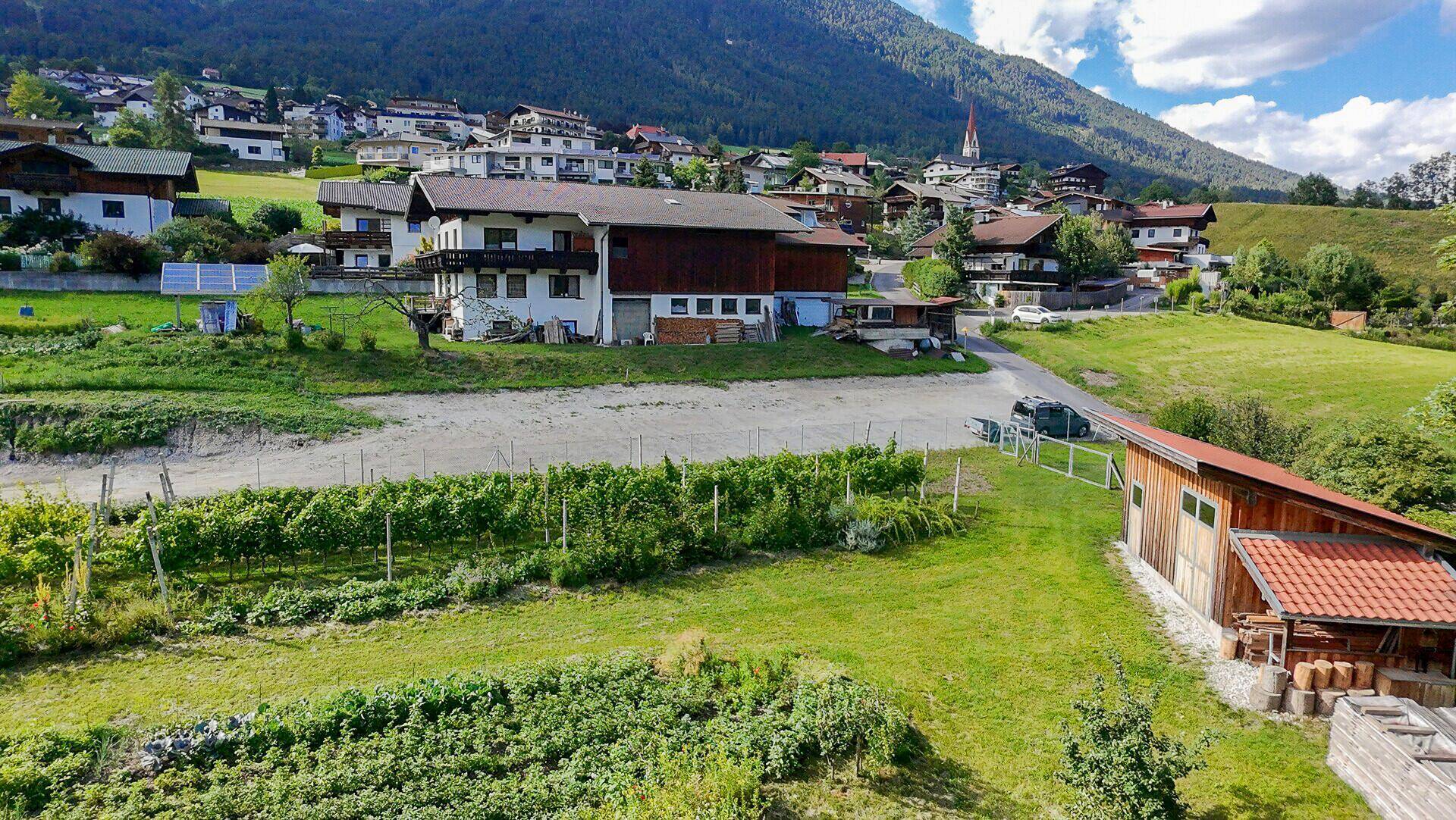 Luftaufnahme einer ländlichen Siedlung mit Häusern, Weinbergen und Feldern vor einer Bergkulisse.