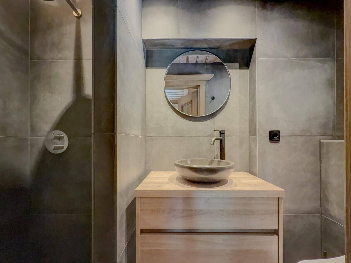 Modernes Badezimmer mit dunklen Fliesen, Holzwaschtisch und rundem Spiegel.